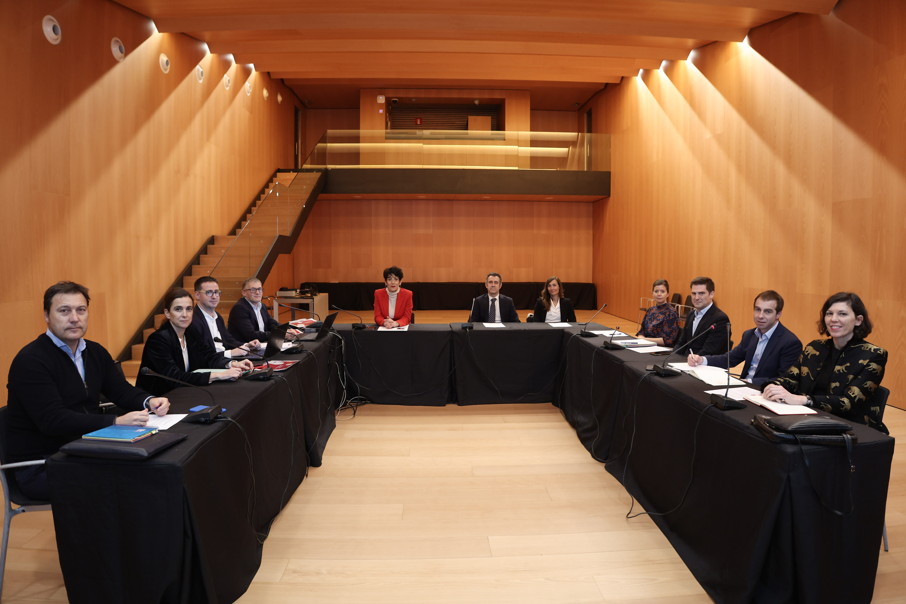 Fotografía frontal de una mesa dispuesta en U, en la que se ve a 11 personas sentadas mirando a cámara en una sala de reuniones