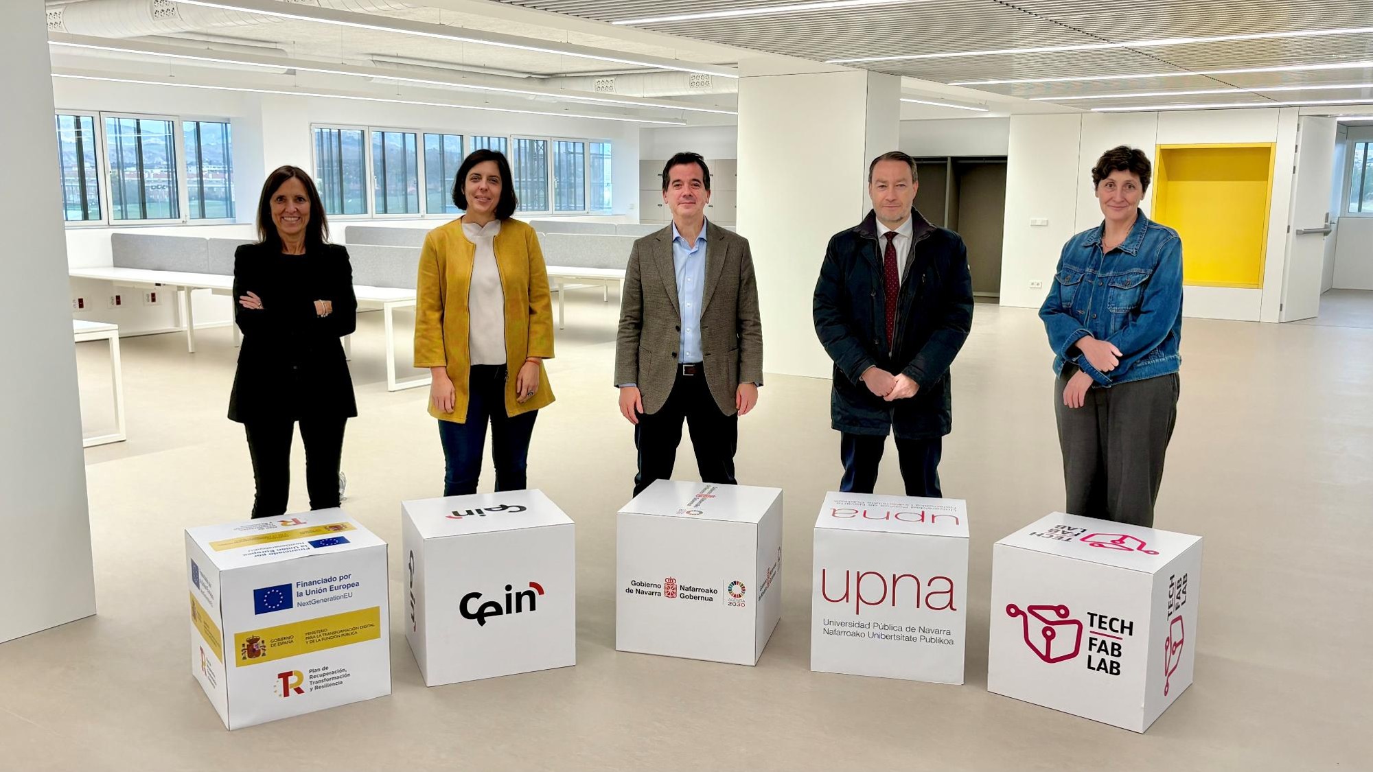 Fotografía de 5 personas mirando a cámara, de pie, en medio de una sala de oficinas de fondo. 
