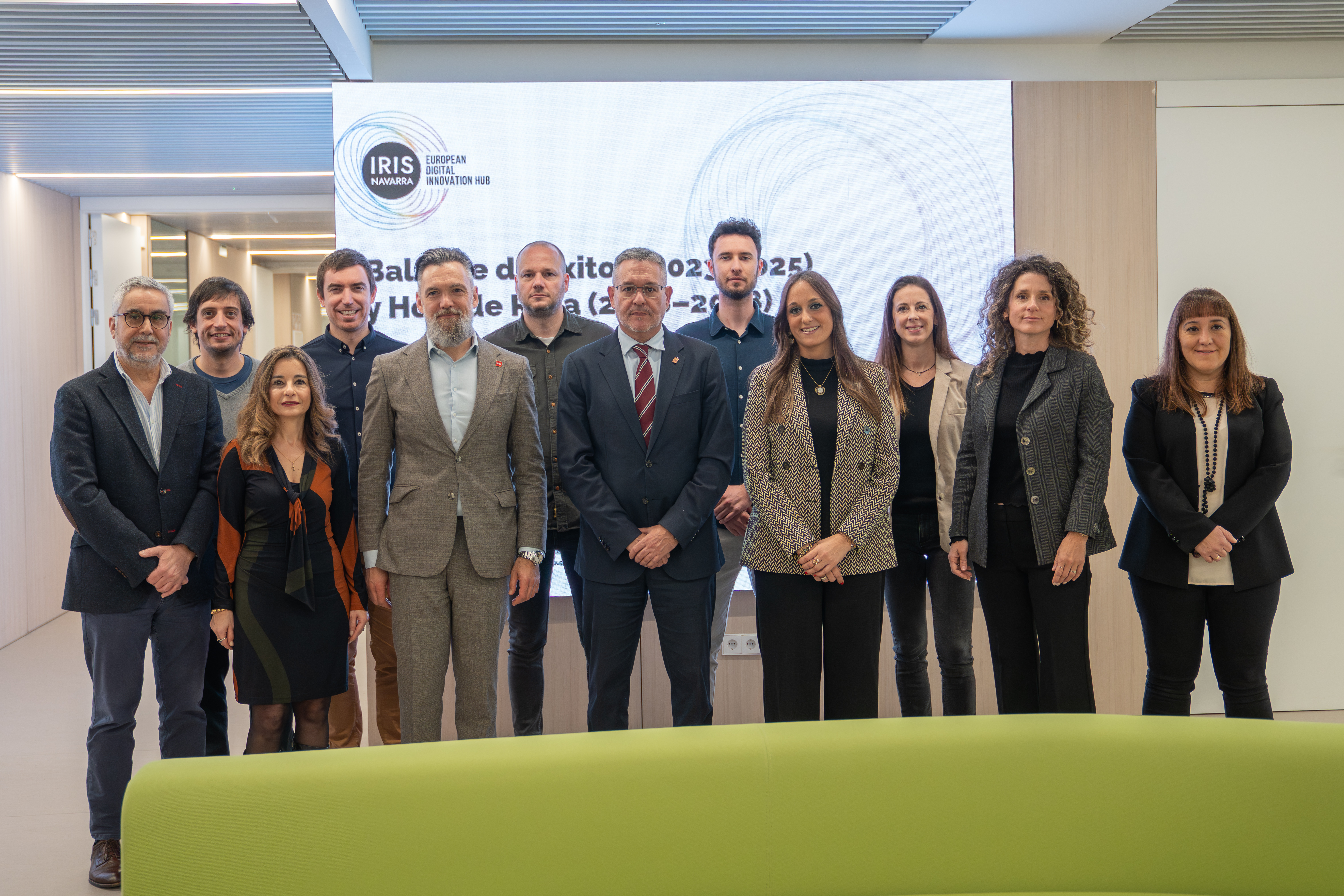 Fotografía de 12 personas mirando a cámara, 7 hombres y 5 mujeres con una pantalla grande detrás con el logotipo de IRIS Navarra