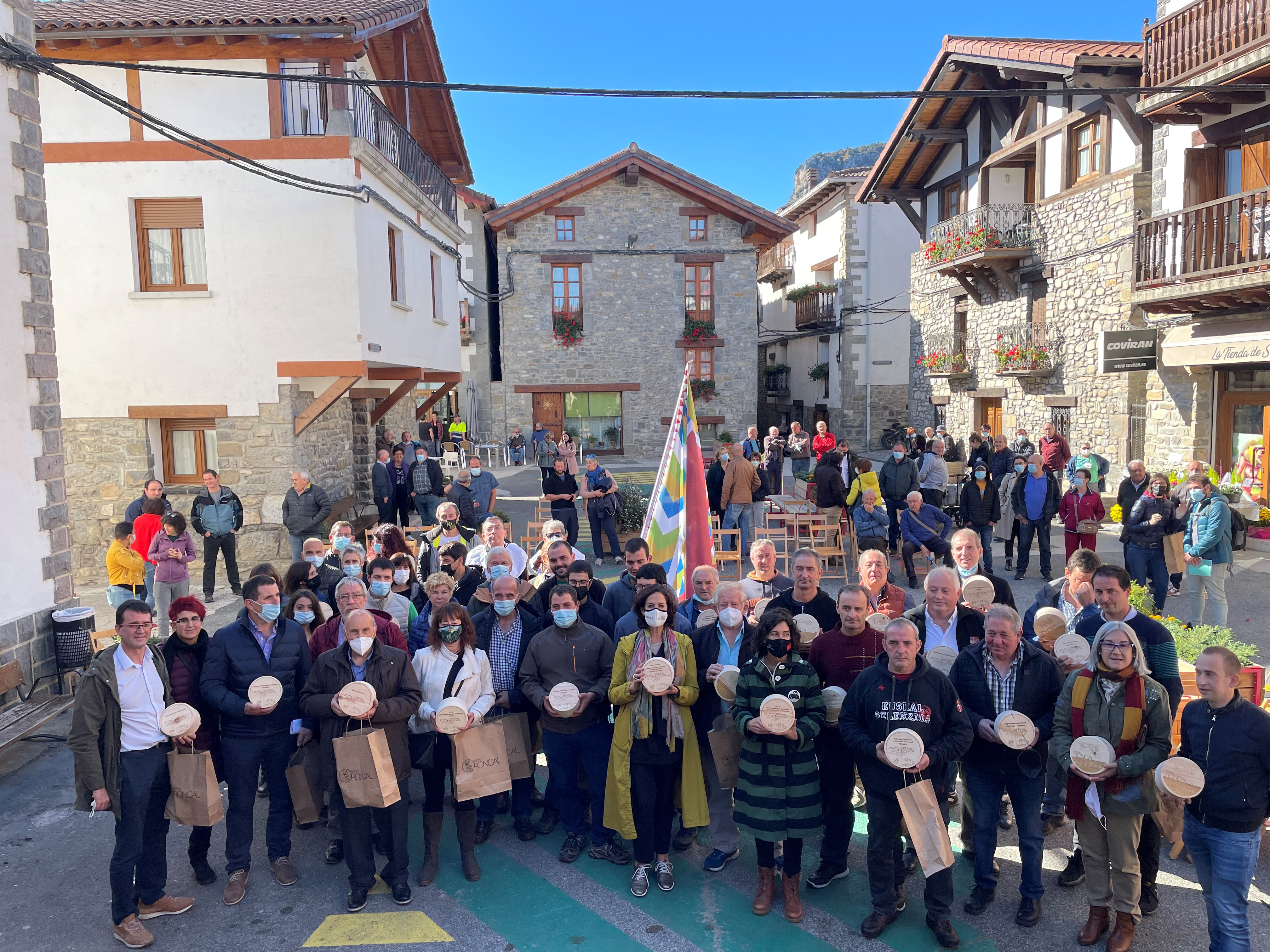Fotografía exterior donde varias personas están de pie en una plaza con un queso del Roncal en sus manos.