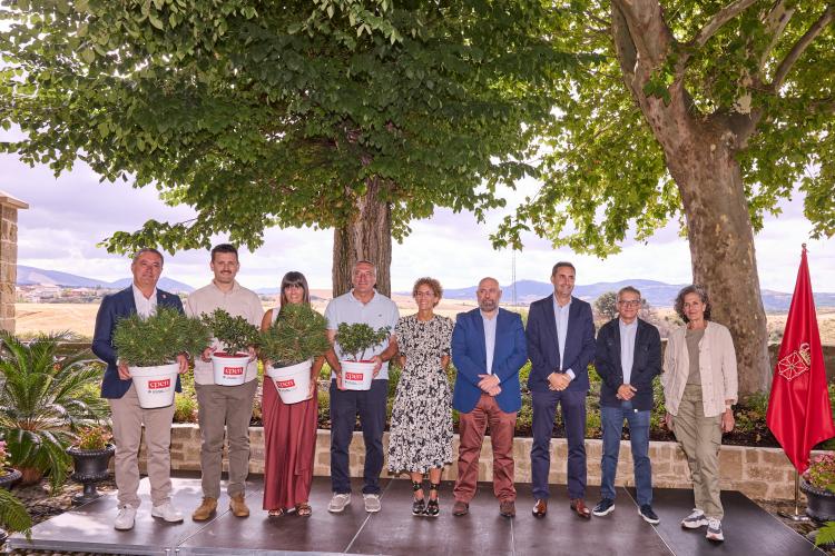9 personas posando en línea en un escenario. 4 de ellas sujetas maceteros con pequeños árboles